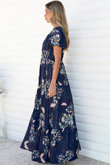 Side view of a woman wearing the Kali Maxi Dress in Colette Print, showing the flutter sleeves, fitted waist and flowing floor-length tiered skirt of the navy floral dress, against a white wall