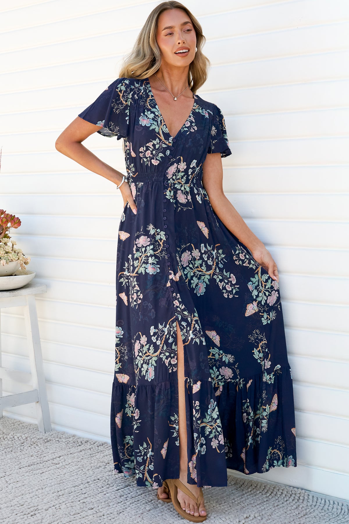 A woman with one hand on her hip wearing the Kali Maxi Dress in Colette Print, a navy floral maxi dress with a V-neck, flutter sleeves, button-front bodice and front split, holding out the tiered skirt against a white wall
