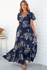A woman walking in the Kali Maxi Dress in Colette Print, with the tiered navy floral skirt swinging out and the front split opening, showing the V-neck, flutter sleeves and button-front bodice, against a white wall
