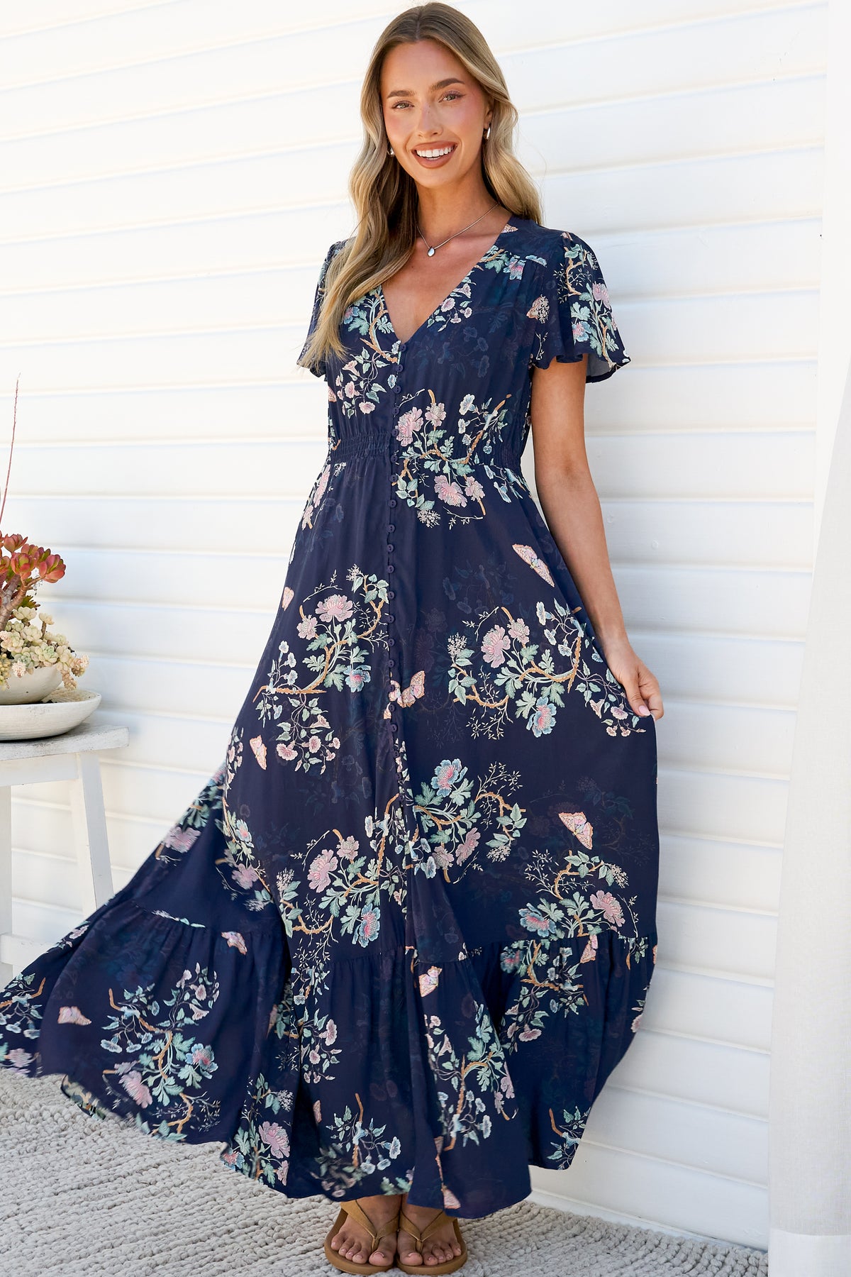 A smiling woman swishing the full tiered skirt of the Kali Maxi Dress in Colette Print, a navy floral maxi dress with a V-neck, flutter sleeves and button-front bodice, against a white wall