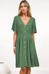 A woman in a green mini dress with a V-neckline, flutter sleeves, and decorative button placket, posed against a white painted brick wall.