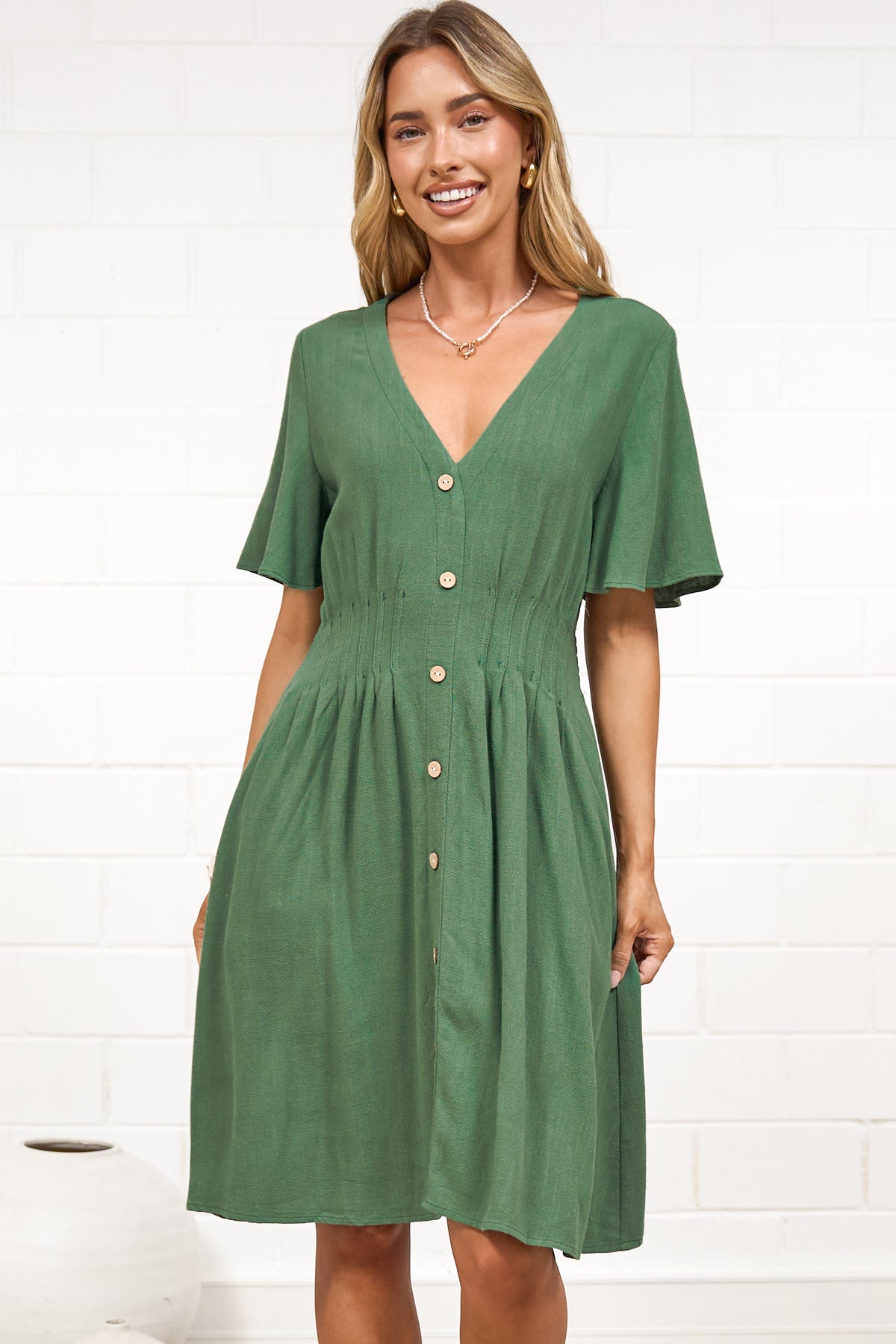A woman in a green mini dress with a V-neckline, flutter sleeves, and decorative button placket, posed against a white painted brick wall.