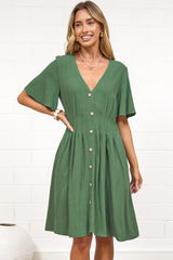 A woman in a green mini dress with a V-neckline, flutter sleeves, and decorative button placket, posed against a white painted brick wall.