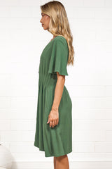 A woman in a green mini dress with a V-neckline, flutter sleeves, and decorative button placket, posed against a white painted brick wall.