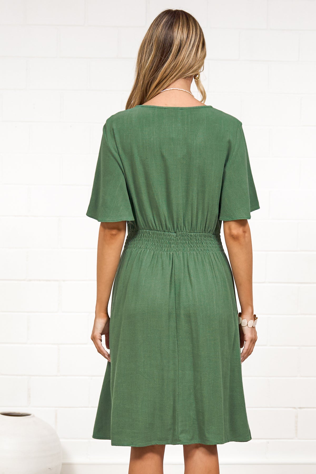 A woman in a green mini dress with a V-neckline, flutter sleeves, and decorative button placket, posed against a white painted brick wall.