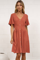 A woman stands in a bright room wearing a short-sleeve terracotta-colored button-up dress and flat  sandals, positioned in front of a white brick wall.