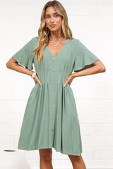 A woman in a sage mini dress with flutter sleeves, a V-neckline, and decorative button placket, posed against a white painted brick wall.