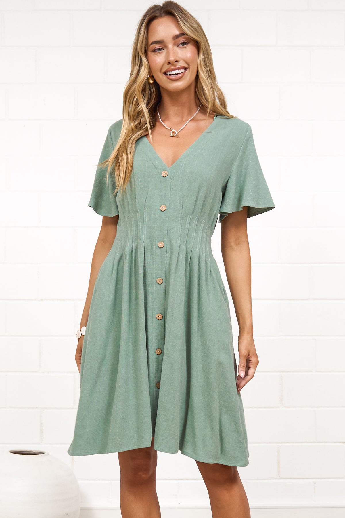 A woman in a sage mini dress with flutter sleeves, a V-neckline, and decorative button placket, posed against a white painted brick wall.
