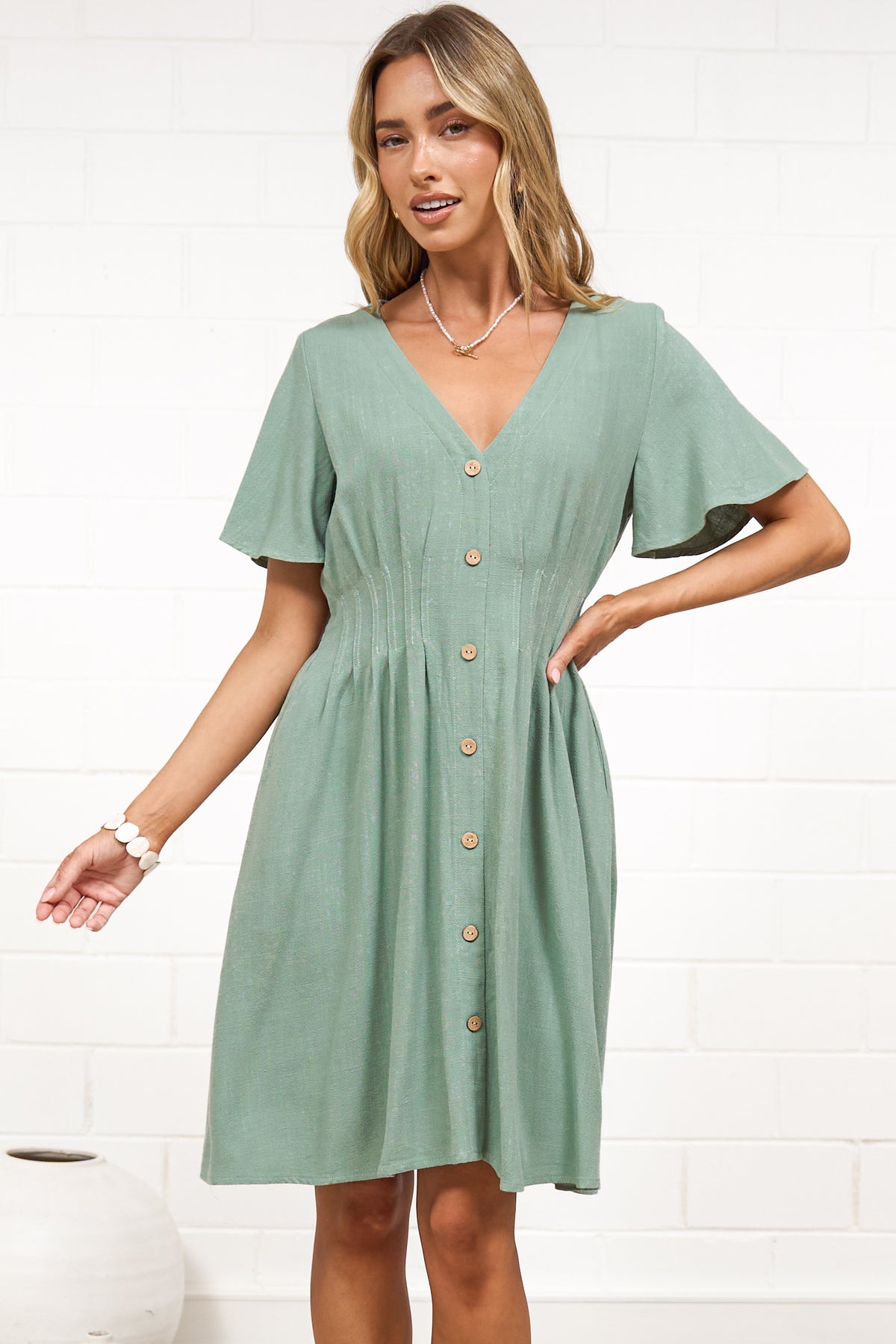 A woman in a sage mini dress with flutter sleeves, a V-neckline, and decorative button placket, posed against a white painted brick wall.