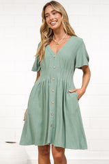 A woman in a sage mini dress with flutter sleeves, a V-neckline, and decorative button placket, posed against a white painted brick wall.