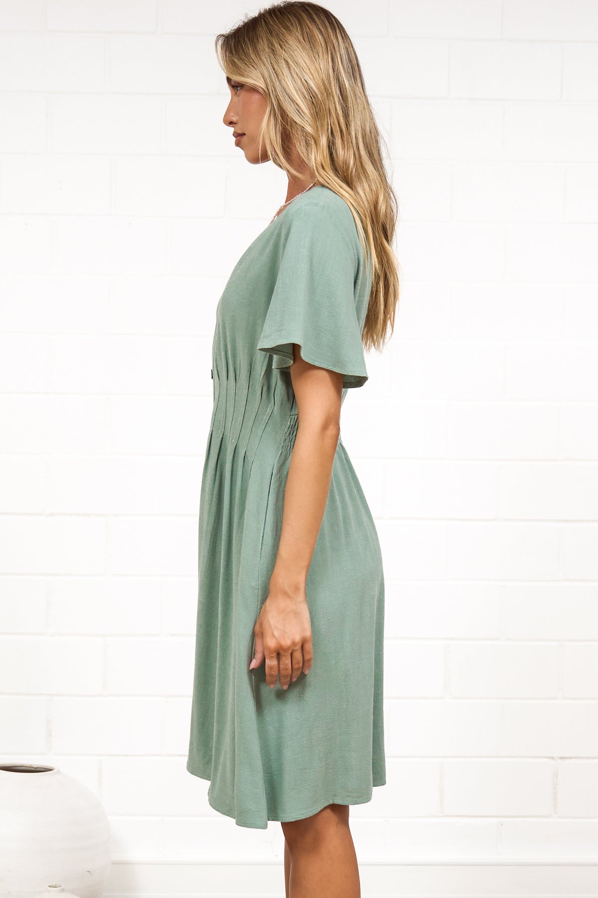 A woman in a sage mini dress with flutter sleeves, a V-neckline, and decorative button placket, posed against a white painted brick wall.