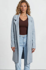 Woman wearing a light blue coat, brown top, and jeans on a plain background