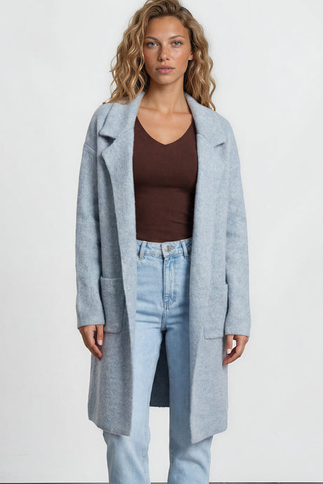 Woman wearing a light blue coat, brown top, and jeans on a plain background