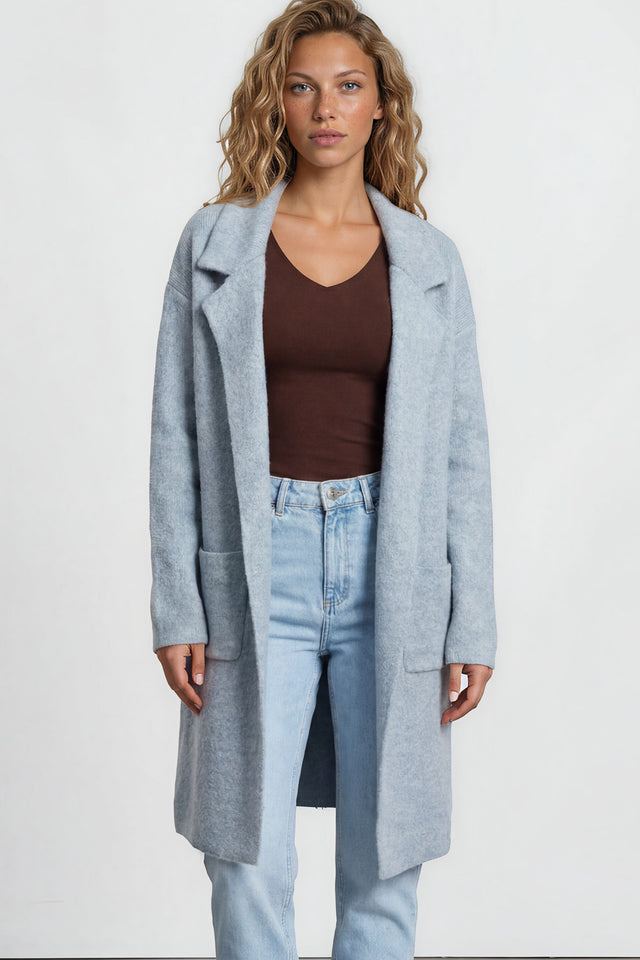 Woman wearing a light blue coat, brown top, and jeans on a plain background