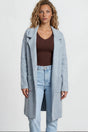 Woman wearing a light blue coat, brown top, and jeans on a plain background