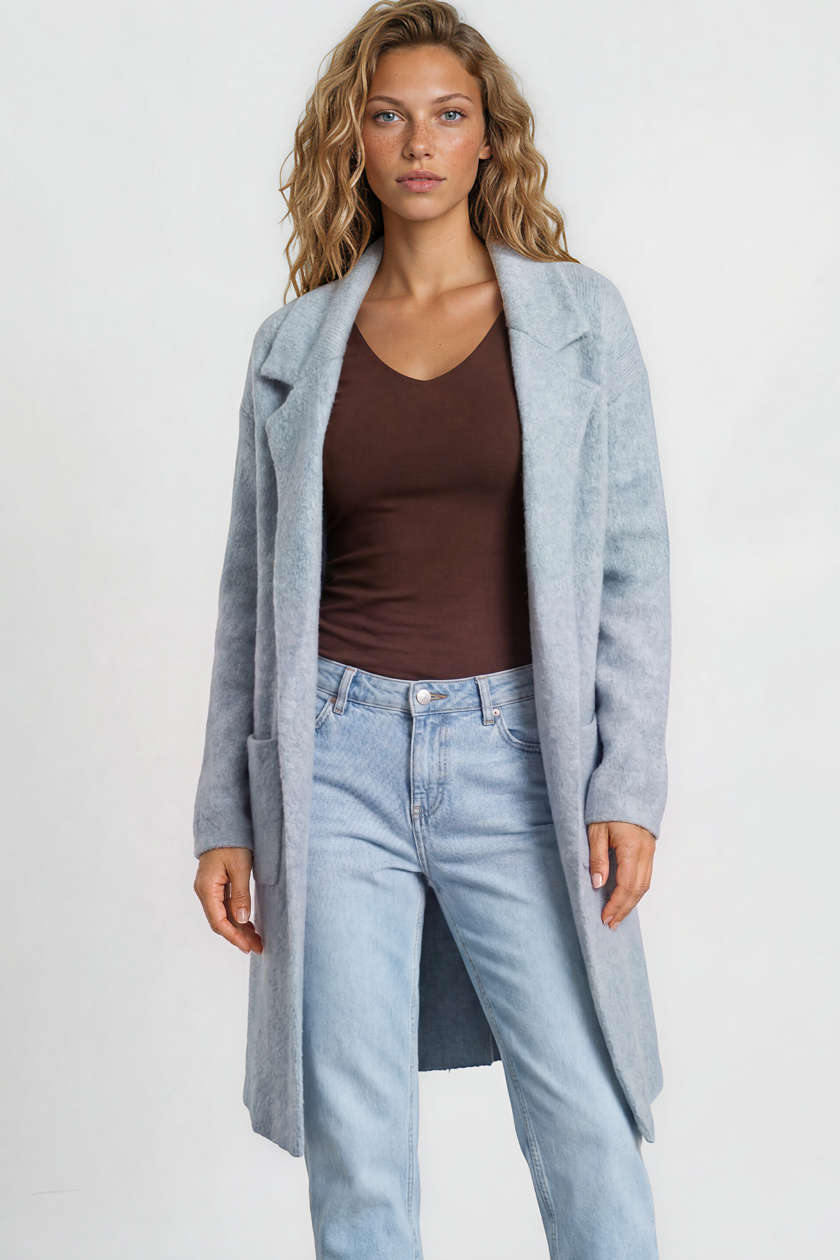 Woman wearing a long light blue coat, brown top, light blue jeans, and white sneakers on a plain background