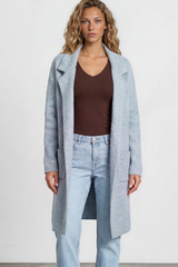 Woman wearing a long light blue coat over a brown top and jeans on a white background