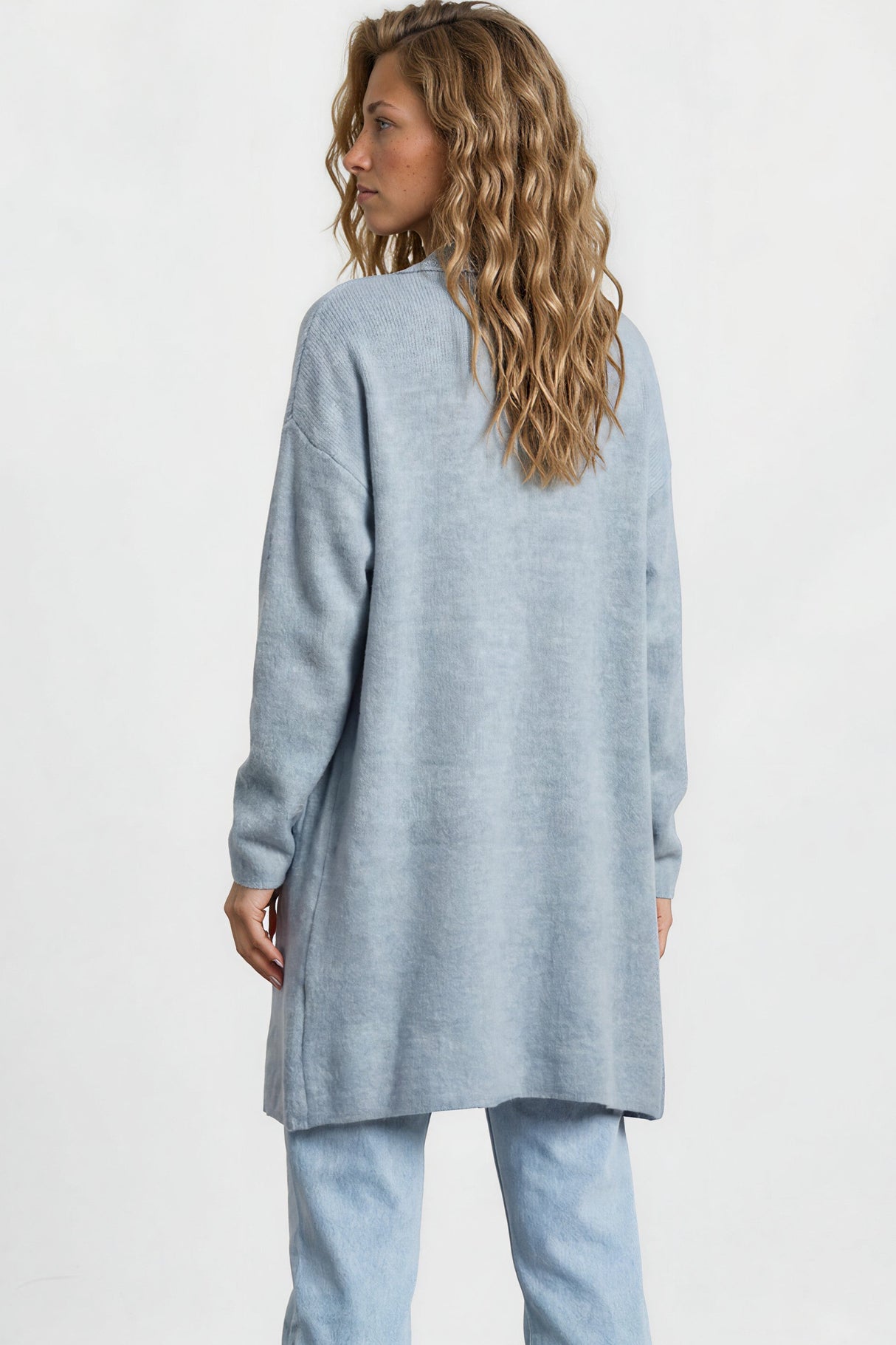 Woman wearing a light blue sweater and jeans standing against a white wall.
