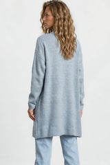 Woman wearing a light blue sweater and jeans standing against a white wall.