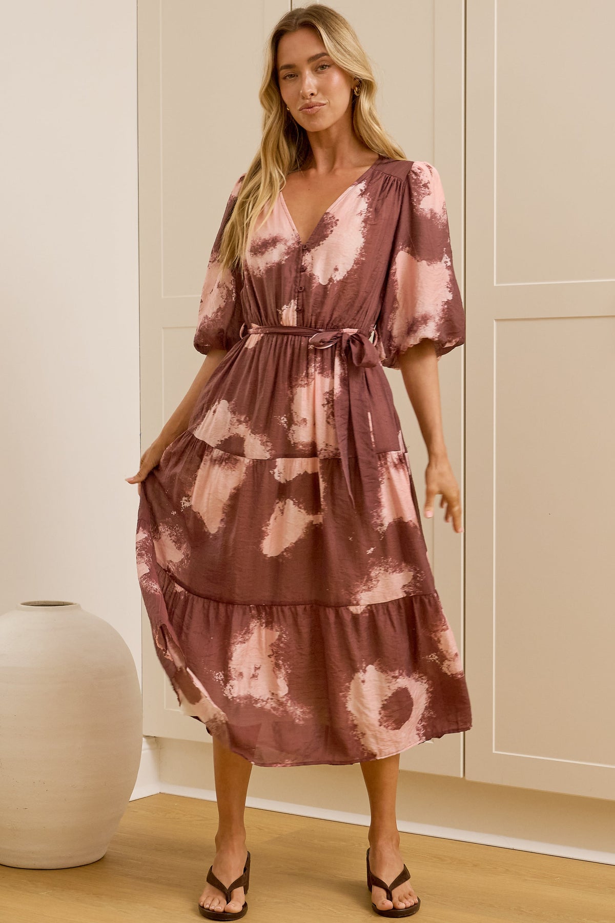 A woman in a flowing printed midi dress featuring balloon sleeves, a partial button front, and adjustable waist tie, posed in front of a warm beige wardrobe.