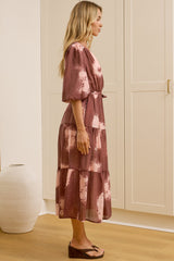 A woman in a flowing printed midi dress featuring balloon sleeves, a partial button front, and adjustable waist tie, posed in front of a warm beige wardrobe.