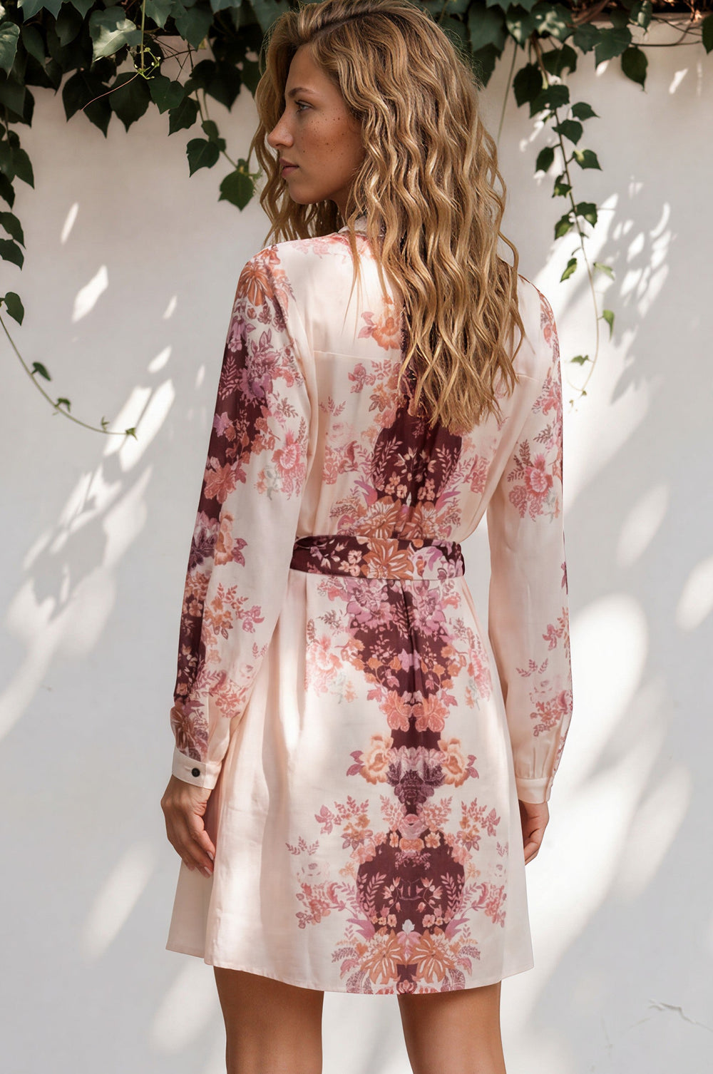 Woman wearing a floral dress standing against a white wall with greenery.