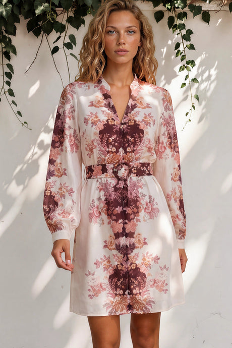 Woman wearing a floral dress standing against a white wall with greenery.