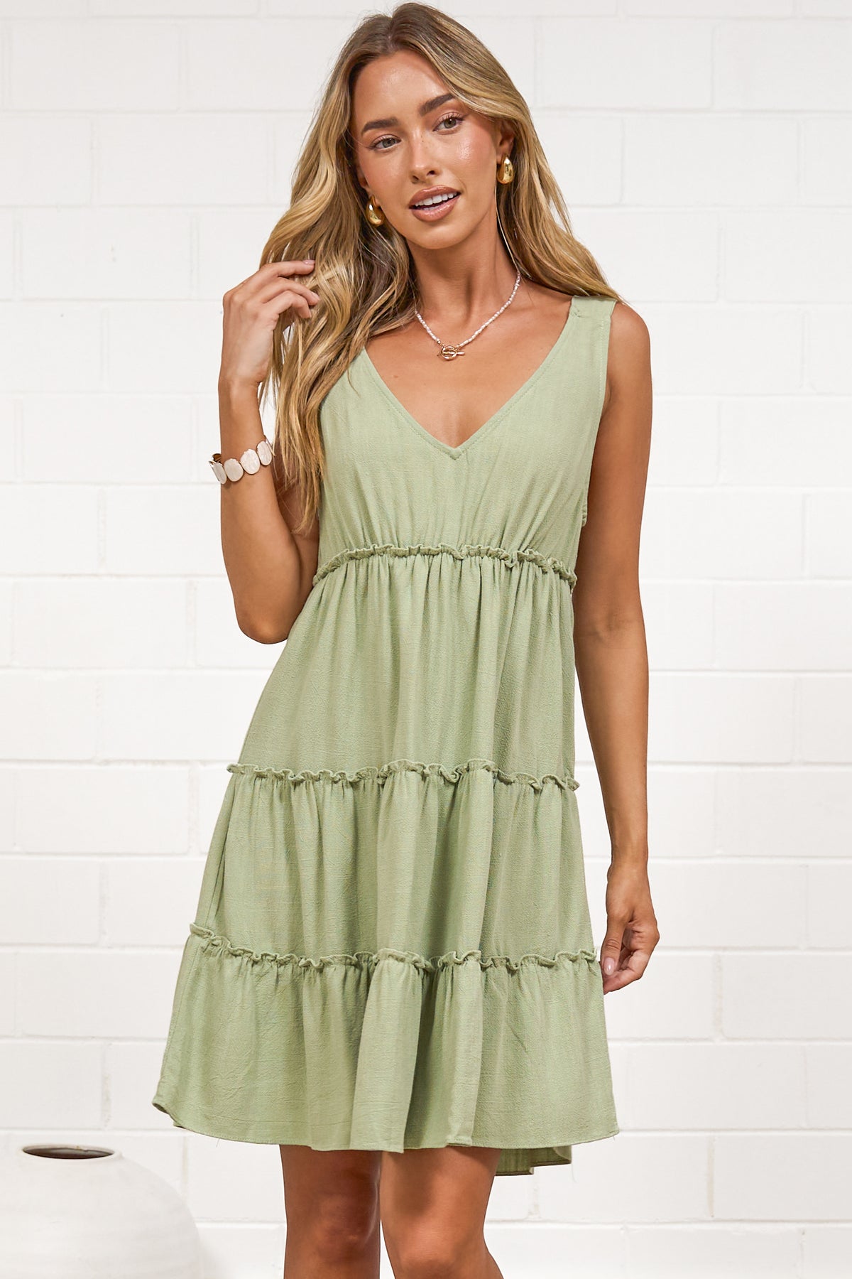 A woman wearing a sage mini dress with tie shoulder straps, a soft V-neckline, tiered skirt, and frill detailing, standing against a white painted brick wall.