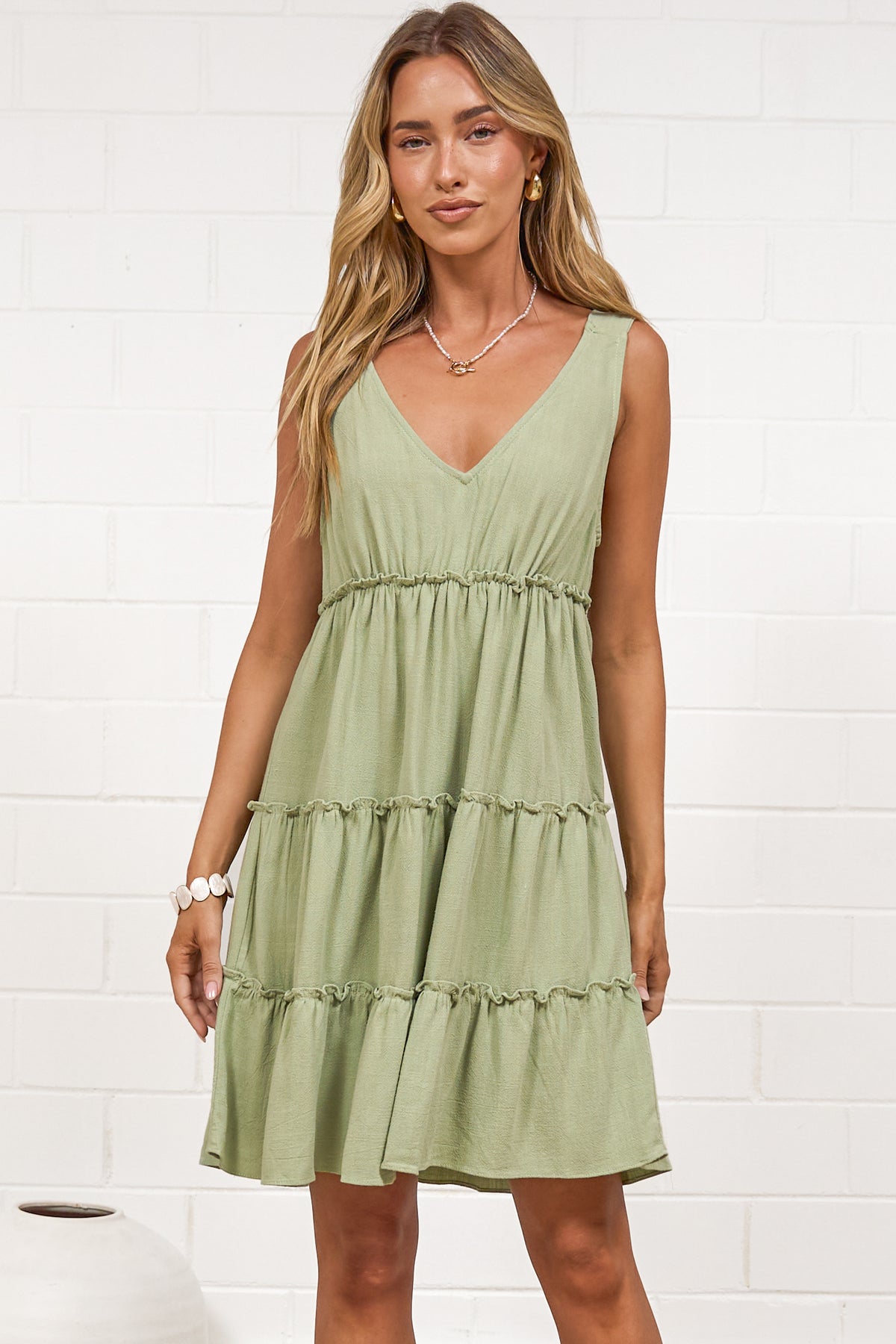 A woman wearing a sage mini dress with tie shoulder straps, a soft V-neckline, tiered skirt, and frill detailing, standing against a white painted brick wall.