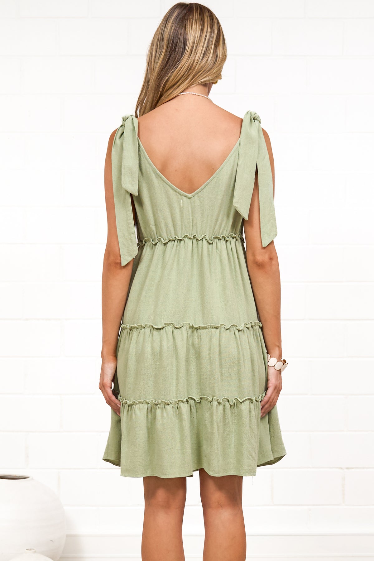 A woman wearing a sage mini dress with tie shoulder straps, a soft V-neckline, tiered skirt, and frill detailing, standing against a white painted brick wall.