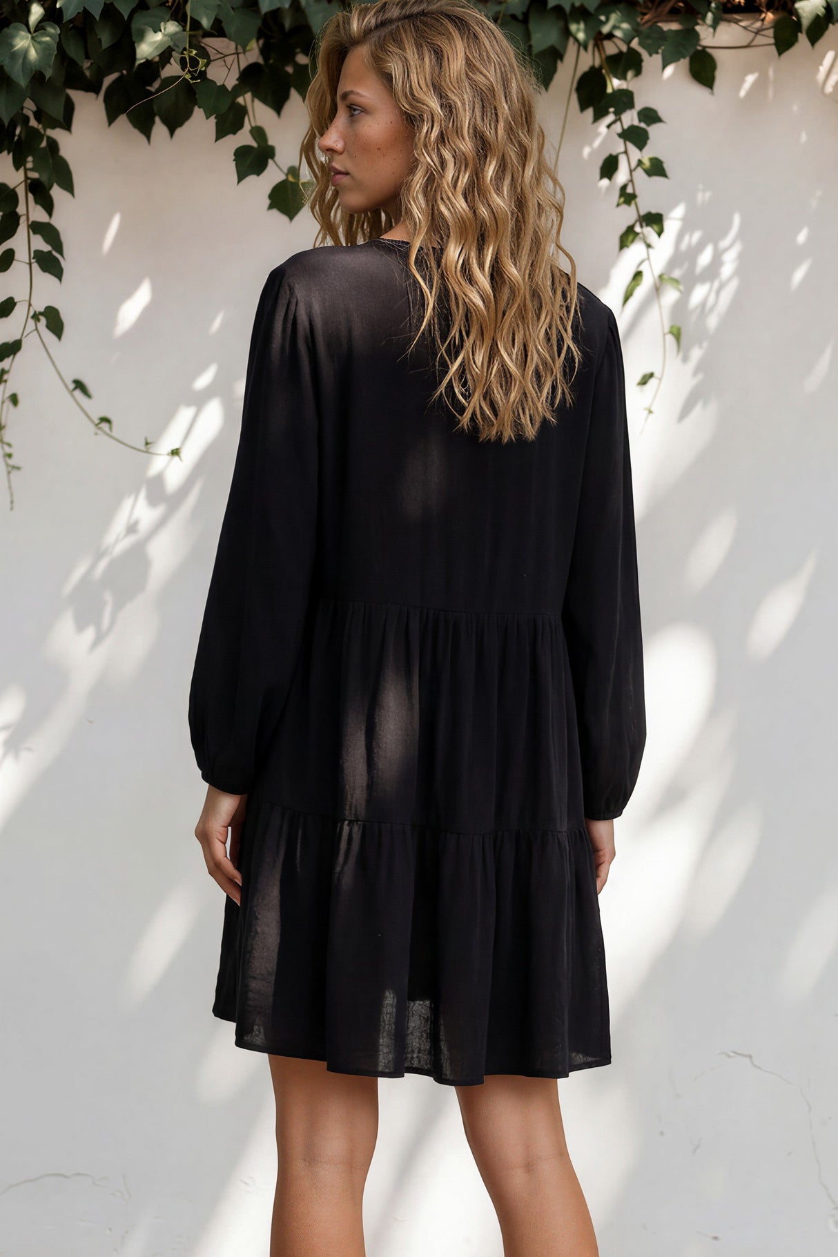 Woman wearing a black dress standing against a white wall with greenery.
