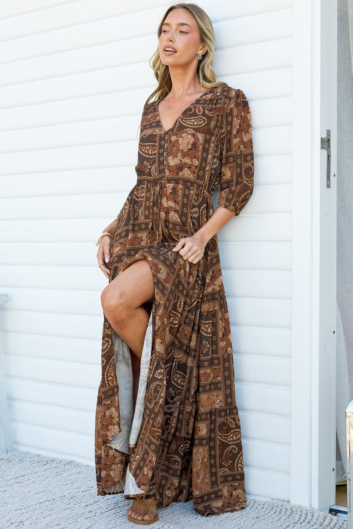 A woman leaning against a white wall in the Lenna Maxi Dress in Mahalla Print, lifting the skirt to show the front split of the brown paisley maxi dress with a V-neck, button front and 3/4 sleeves