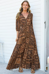 A smiling woman walking in the Lenna Maxi Dress in Mahalla Print, a V-neck button-front maxi dress with 3/4 sleeves, a tie waist and tiered skirt in a brown and rust bohemian paisley print, against a white wall