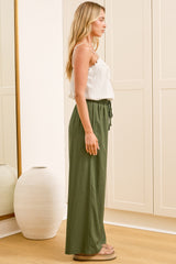 A woman wearing khaki linen pants with an elasticated waist, tie detail, and straight-leg fit, standing in front of a warm beige closet.