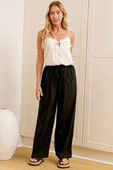 A woman wearing black linen pants with an elasticated waist, tie detail, and straight-leg fit, standing in front of a warm beige closet.