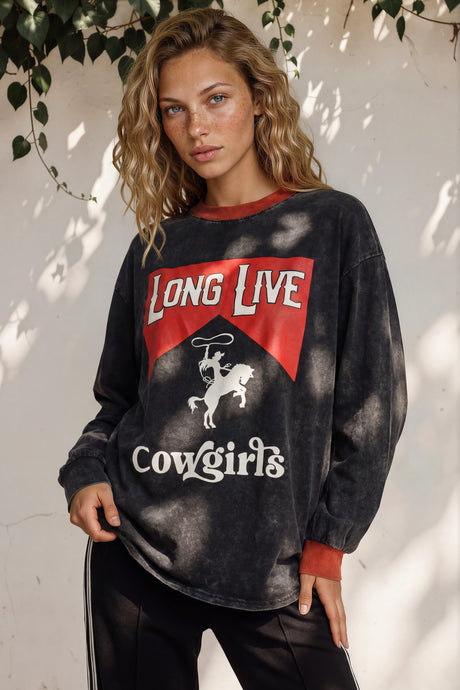 Woman wearing a sweatshirt with 'Long Live Cowgirls' text and design, standing against a white wall with greenery.