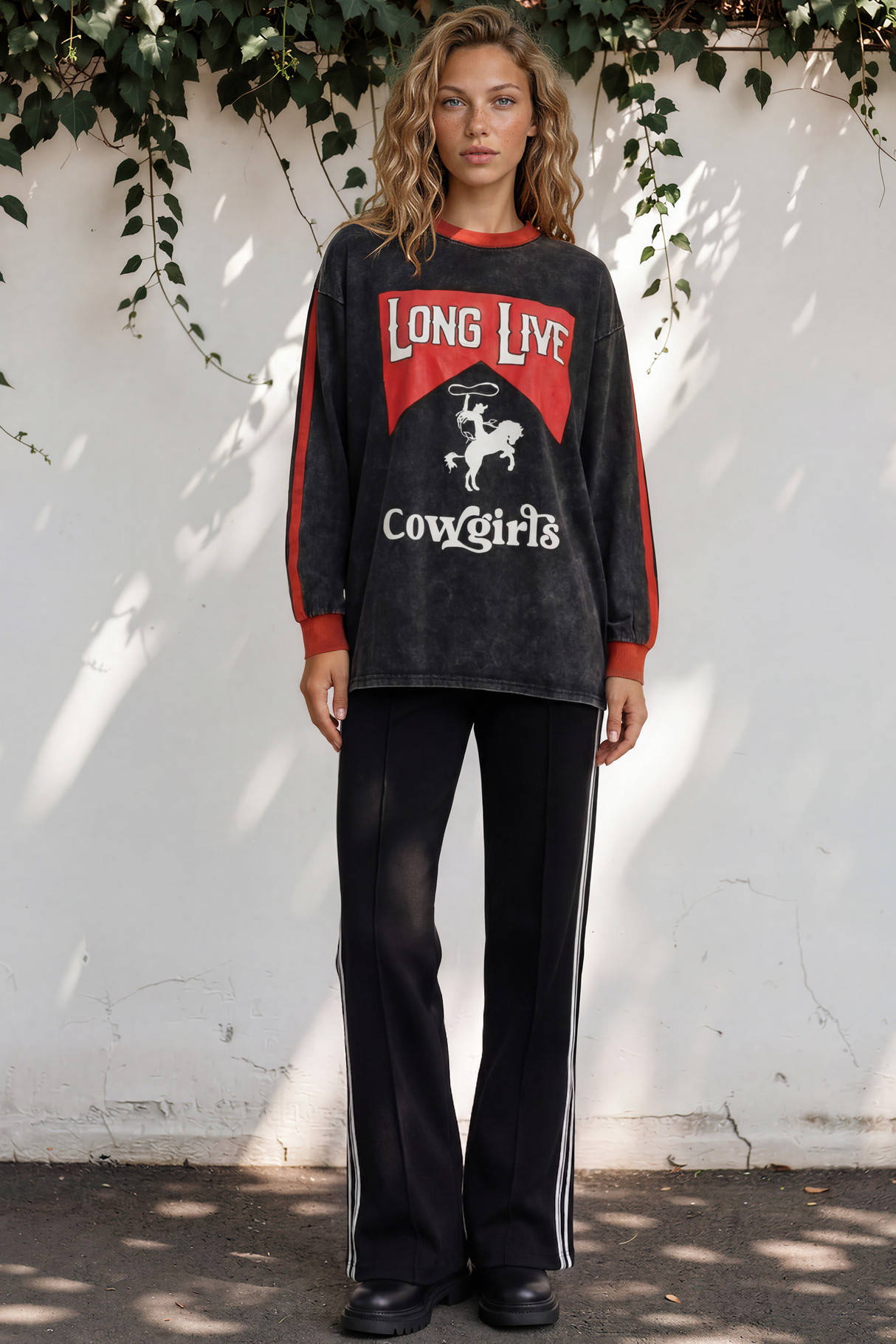 Woman wearing a black and red long-sleeve shirt with 'Long Live Cowgirls' text, standing against a white wall with greenery.