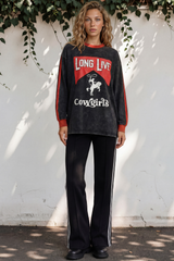Woman wearing a black and red long-sleeve shirt with 'Long Live Cowgirls' text, standing against a white wall with greenery.