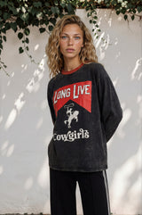 Woman wearing a black sweatshirt with 'Long Live Cowgirls' text and graphics against a white wall.
