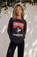 Woman wearing a black sweatshirt with red and white text and graphics, standing against a white wall with greenery.