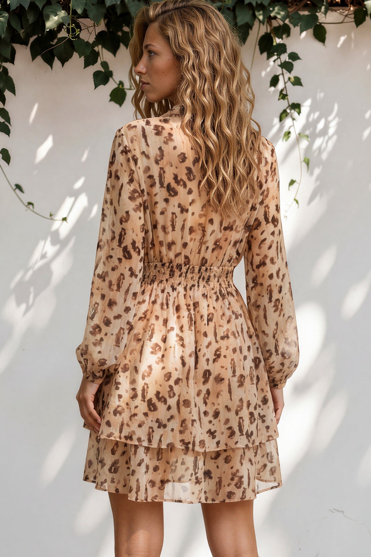 Woman wearing a patterned dress standing against a white wall with greenery.