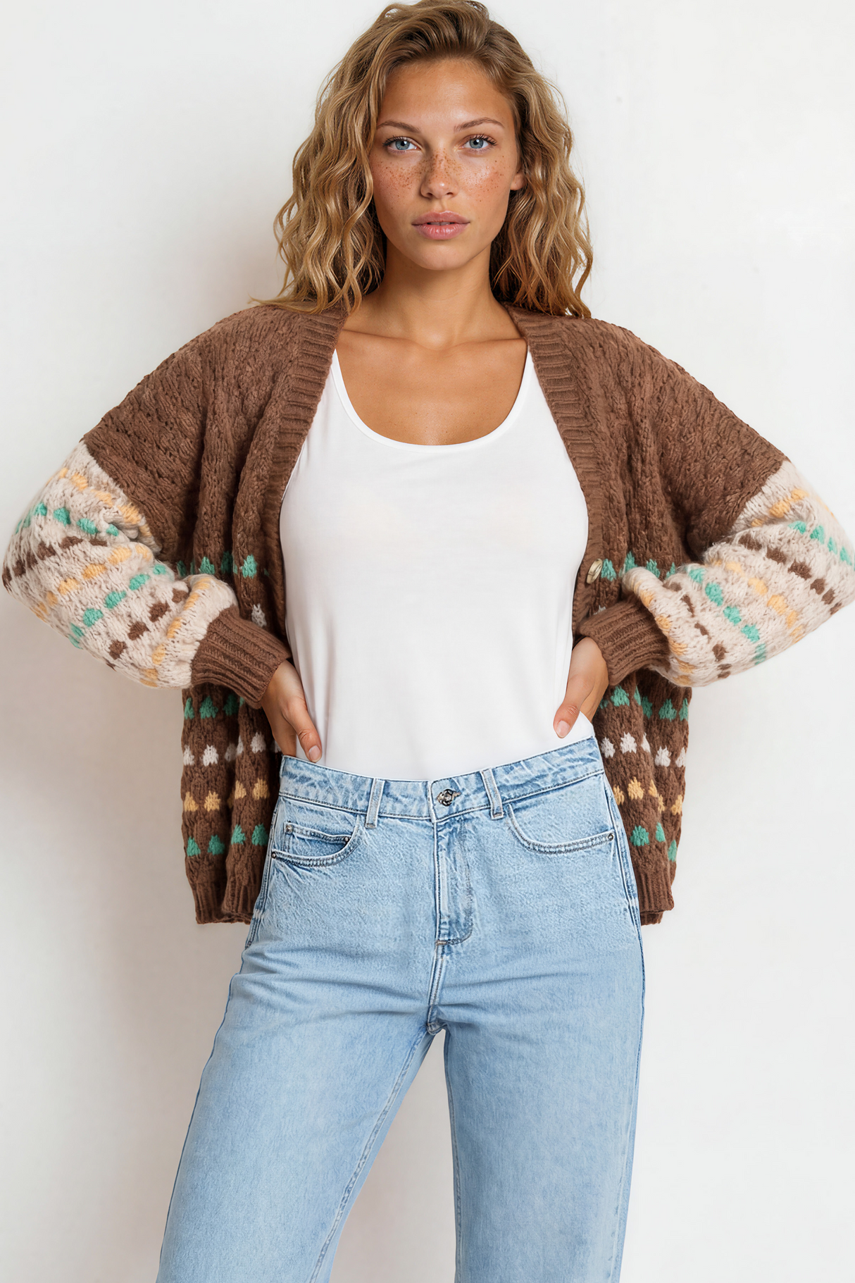 Woman wearing a brown patterned cardigan, white tank top, and light blue jeans against a white background