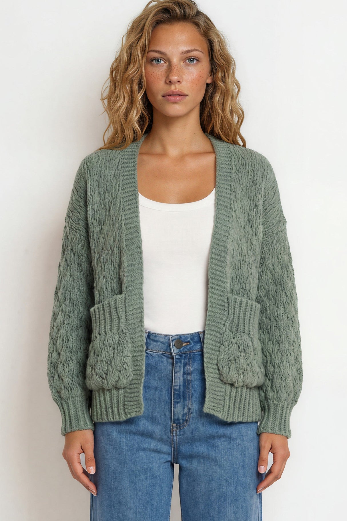 Woman wearing a green cardigan over a white top and blue jeans on a plain background