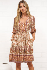 A woman in a tan paisley print mini dress with balloon sleeves, a V-neckline, tie-waist detail, and tiered skirt, posed against a white painted brick wall.
