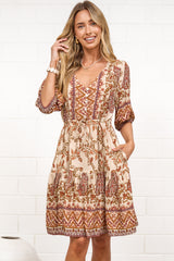 A woman in a tan paisley print mini dress with balloon sleeves, a V-neckline, tie-waist detail, and tiered skirt, posed against a white painted brick wall.