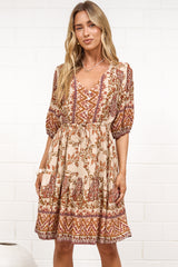 A woman in a tan paisley print mini dress with balloon sleeves, a V-neckline, tie-waist detail, and tiered skirt, posed against a white painted brick wall.