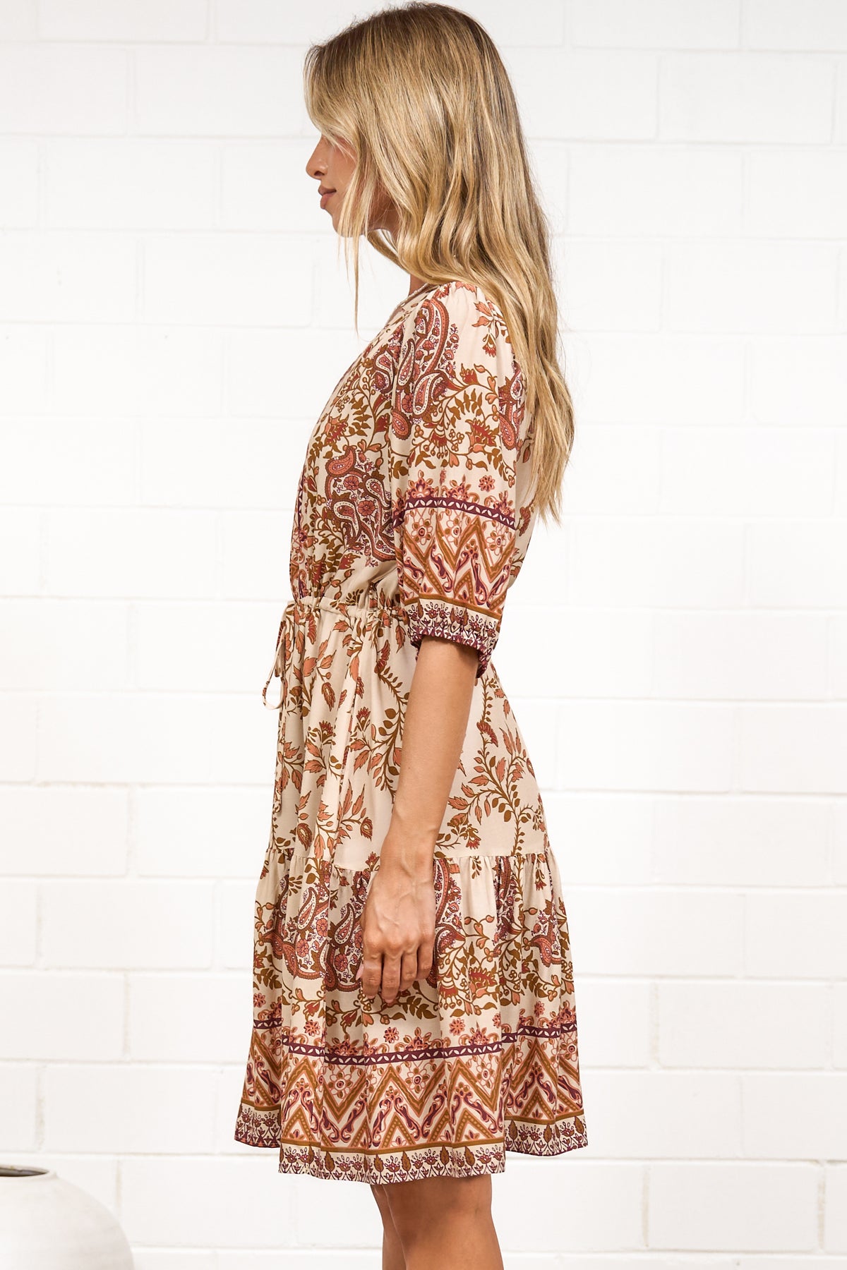 A woman in a tan paisley print mini dress with balloon sleeves, a V-neckline, tie-waist detail, and tiered skirt, posed against a white painted brick wall.