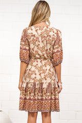 A woman in a tan paisley print mini dress with balloon sleeves, a V-neckline, tie-waist detail, and tiered skirt, posed against a white painted brick wall.
