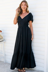A smiling woman with one hand in her hair leaning against a white wall in the Marilyn Maxi Dress in Black, a floor-length dress with a smocked button-front bodice, flutter sleeves and lace-trimmed tiered hem