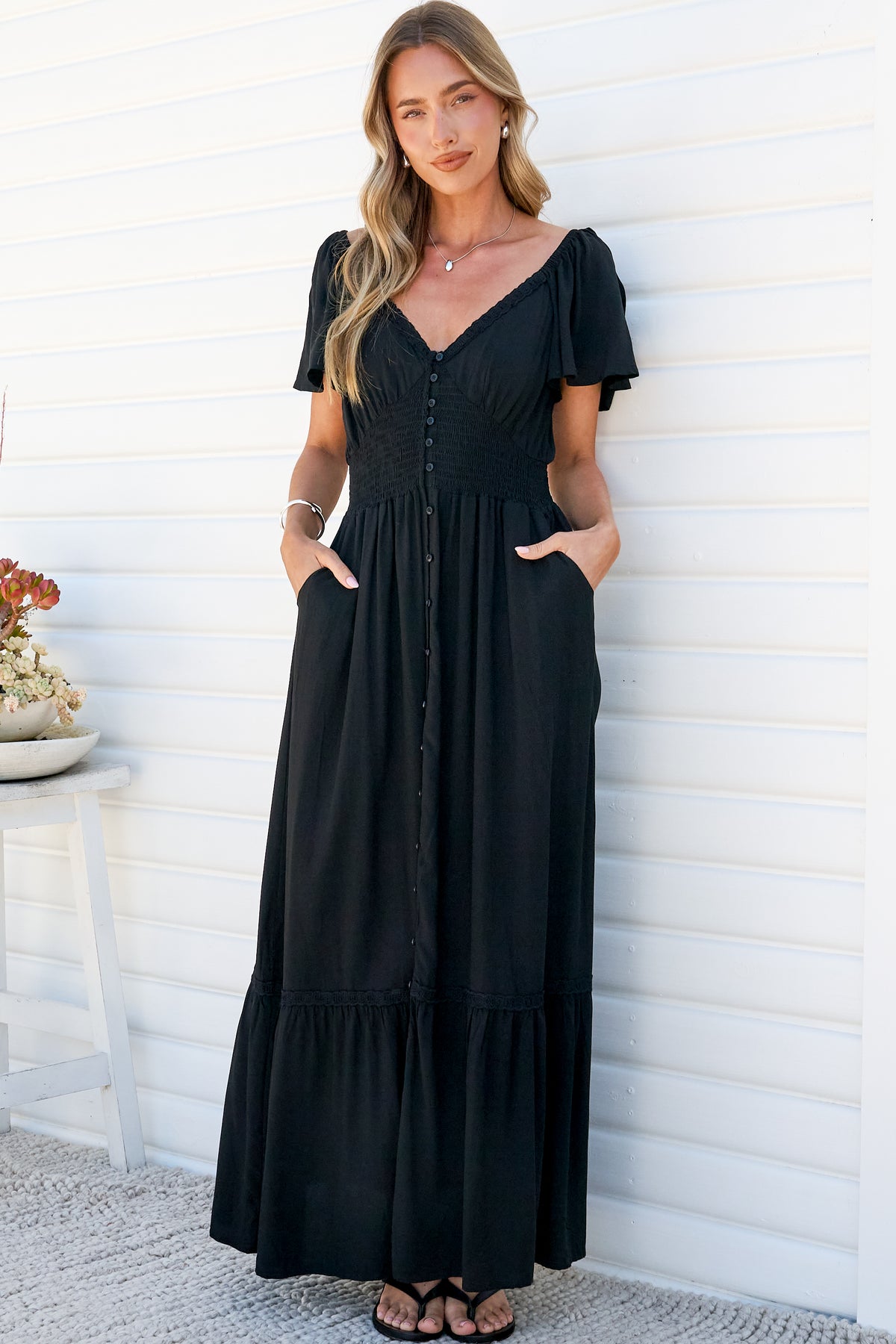 A woman with both hands in her pockets wearing the Marilyn Maxi Dress in Black, a relaxed maxi dress with a V-shaped sweetheart neckline, flutter sleeves, smocked button-front bodice and tiered hem, against a white wall